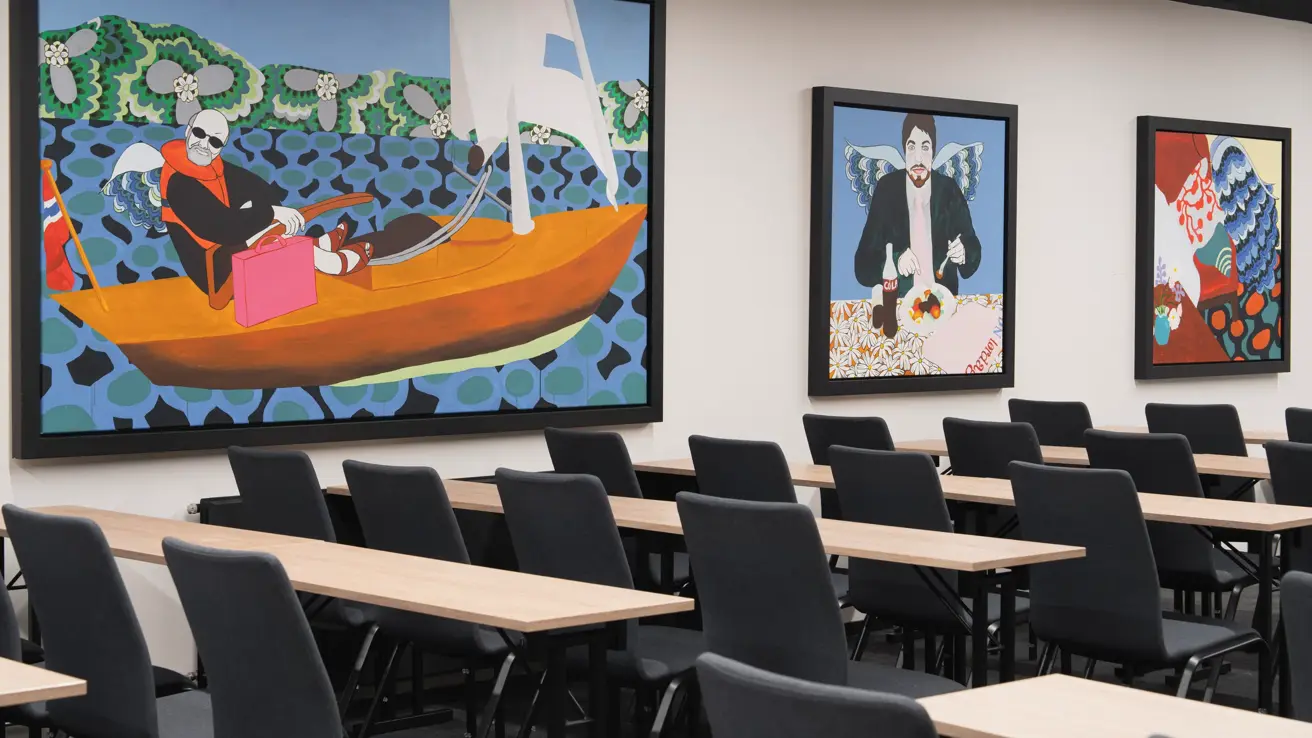 Møterom med klasseroms oppsett, med svarte stoler og lyse trebord. Fargerike bilder på veggene