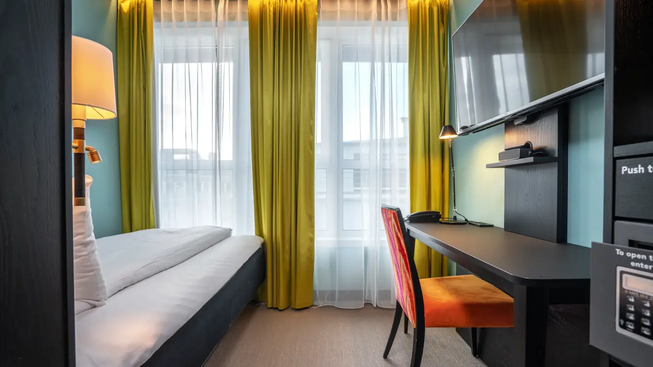 Singelrom med enkeltseng, skrivebord, vegghengt TV, oransje stol og gule gardiner.