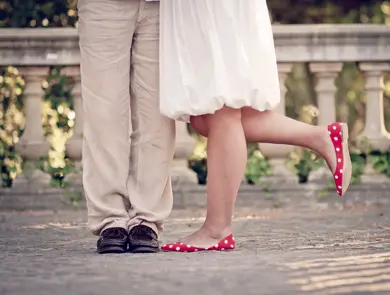 Halvfigur av bein på dame med røde ballerinasko og hvit kjole, og mann med beige dressbukse og mørke skinnsko, som står i et parkområde. Foto.