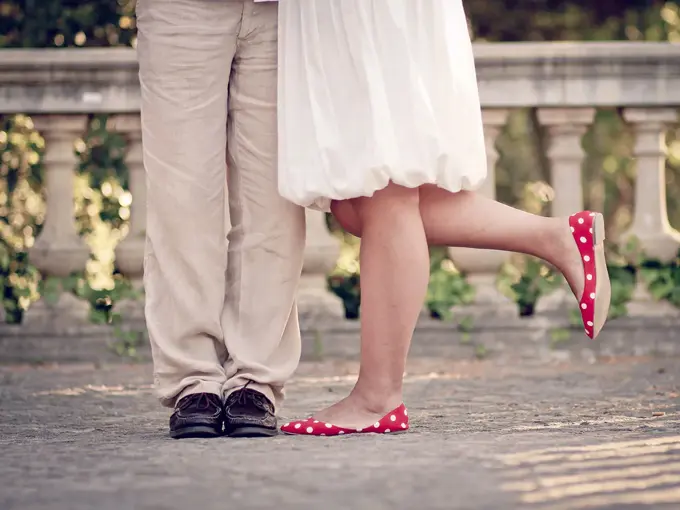 Halvfigur av bein på dame med røde ballerinasko og hvit kjole, og mann med beige dressbukse og mørke skinnsko, som står i et parkområde. Foto.