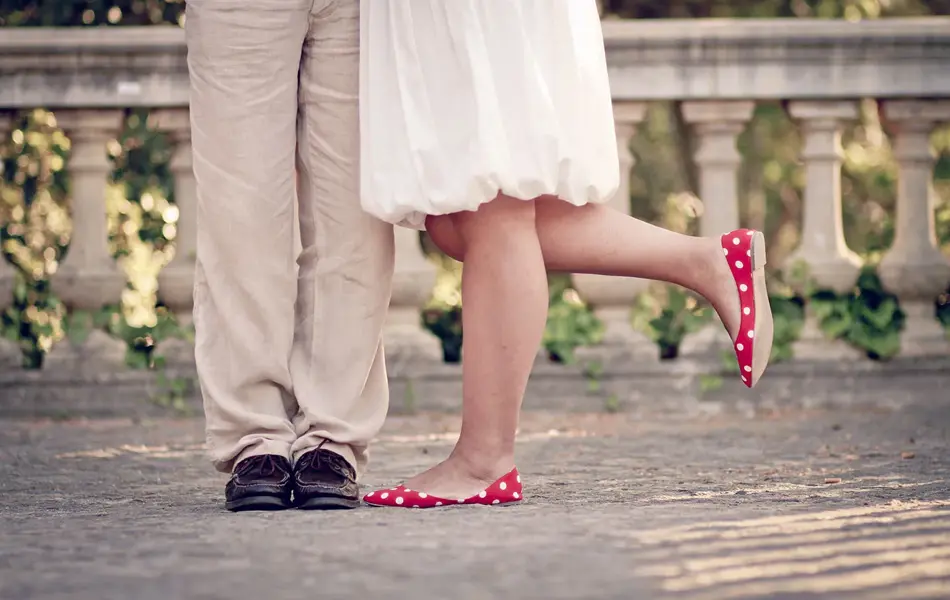 Halvfigur av bein på dame med røde ballerinasko og hvit kjole, og mann med beige dressbukse og mørke skinnsko, som står i et parkområde. Foto.