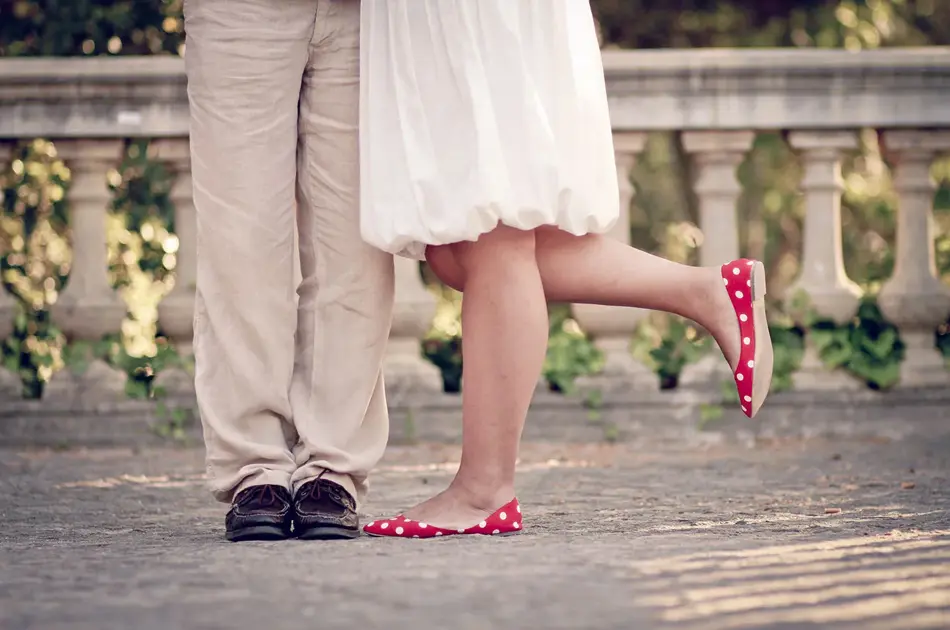 Halvfigur av bein på dame med røde ballerinasko og hvit kjole, og mann med beige dressbukse og mørke skinnsko, som står i et parkområde. Foto.
