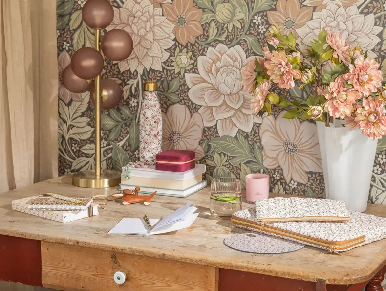 Et skrivebord med notatbøker, en lampe og en vase med blomster.