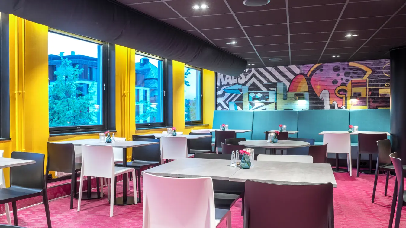 Frokostsal, diverse spisebord, hvite og svarte stoler, rosa gulvteppe, vinduer, gule vegger. 