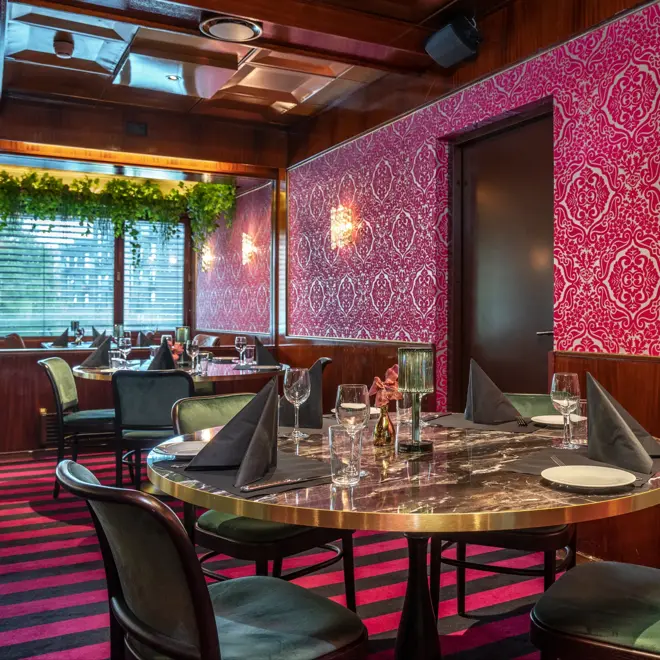 Middagsbord i restaurant, grønne stoler, rosa gulvteppe, rosa tapetvegg med mønster, vindu, runde spisebord
