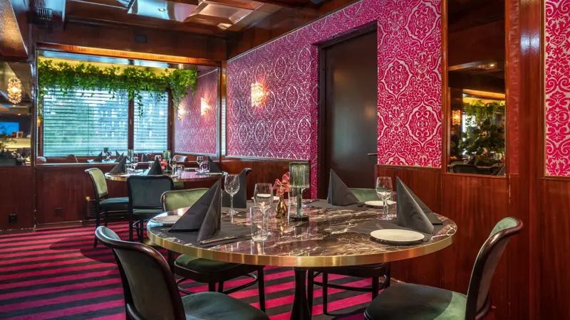 Middagsbord i restaurant, grønne stoler, rosa gulvteppe, rosa tapetvegg med mønster, vindu, runde spisebord