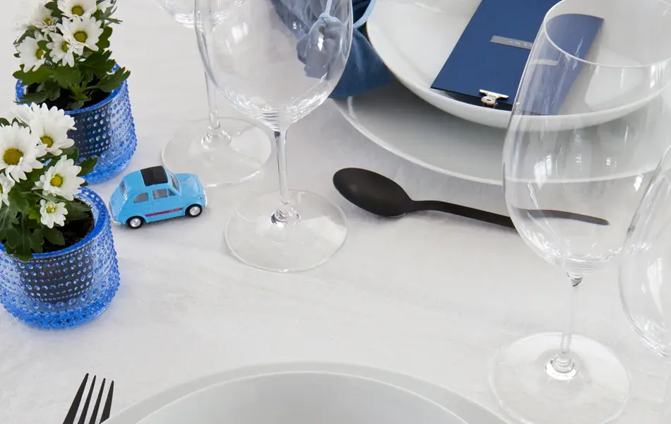 Steg 6 - bord dekket med mørkeblå tøyservietter og blått menykort, hvite blomster, en liten lekebil og tomme vinglass