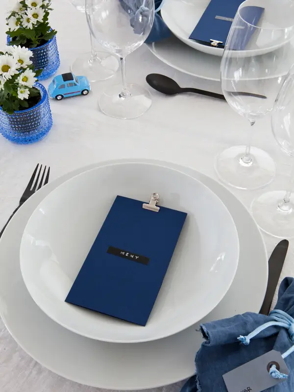 Steg 6 - bord dekket med mørkeblå tøyservietter og blått menykort, hvite blomster, en liten lekebil og tomme vinglass