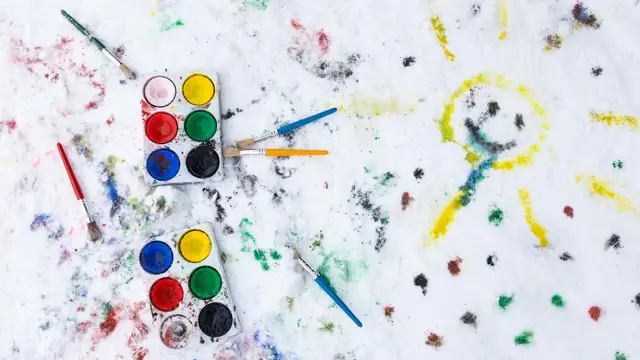 To maleskrin og flere pensler som ligger i snøen, hvor det er malt en smilende sol og prikker i ulike farger