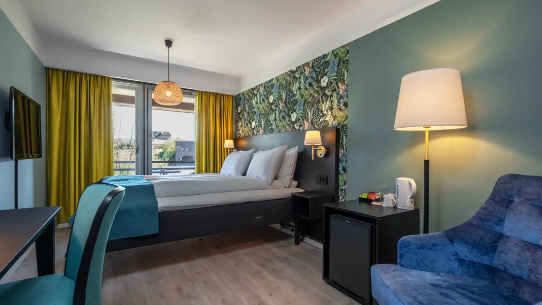 Standardrom med queen seng, blått sengeteppe, mønstrete og fargerik tapet bak sengen, gule gardiner, mørkeblå lenestol, vegghengt TV og skrivebord med tilhørende turkis stol