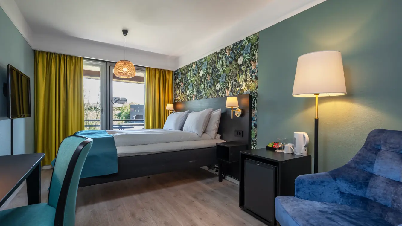 Standardrom med queen seng, blått sengeteppe, mønstrete og fargerik tapet bak sengen, gule gardiner, mørkeblå lenestol, vegghengt TV og skrivebord med tilhørende turkis stol