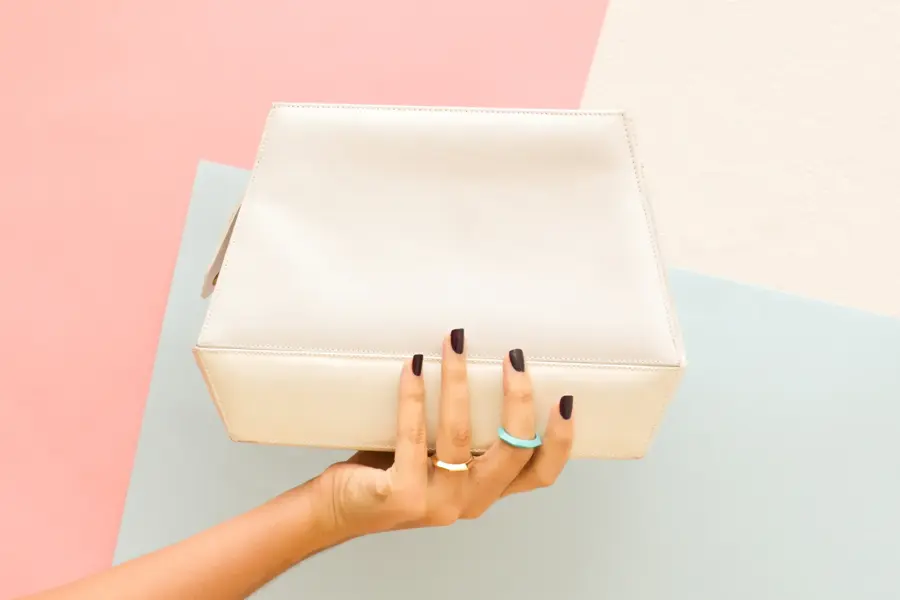 En hånd som holder en hvit håndveske foran farget bakgrunn