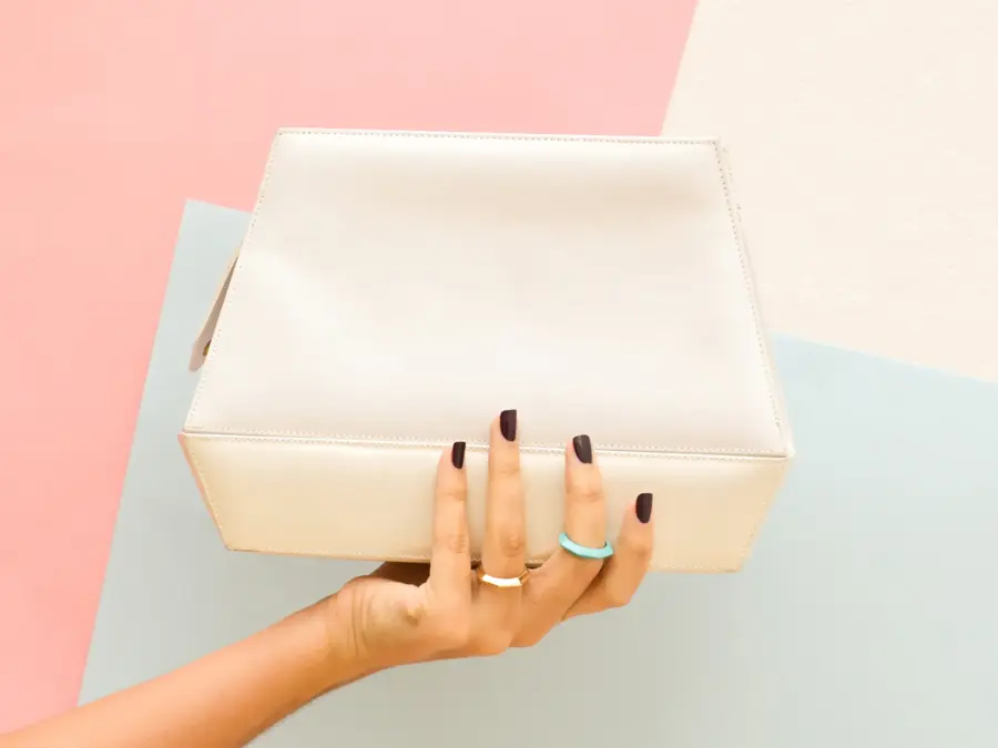 En hånd som holder en hvit håndveske foran farget bakgrunn