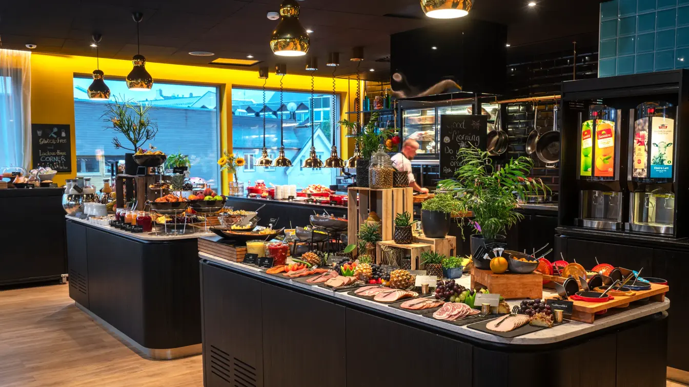 Tre store vinduer fra gulv til tak, gule vegger, svart tak, store bordbenker dandert med  pålegg, frukt og planter