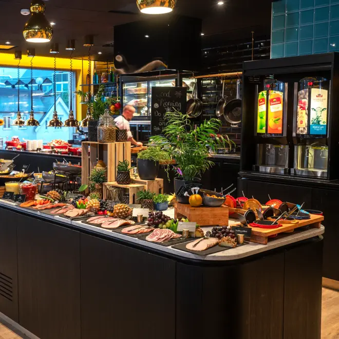 Tre store vinduer fra gulv til tak, gule vegger, svart tak, store bordbenker dandert med  pålegg, frukt og planter