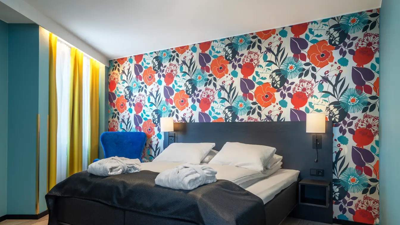 Blomstertapetvegg, dobbeltseng, veggspeil, vindu, gule gardiner, parkettgulv, blå stol 