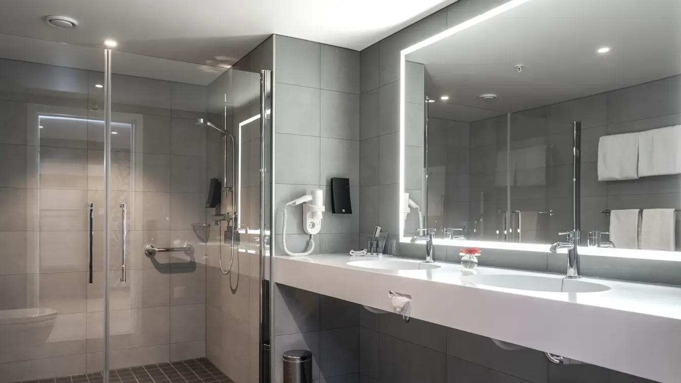 Grå veggfliser, dusj, speil, vask, svarte gulvfliser. 