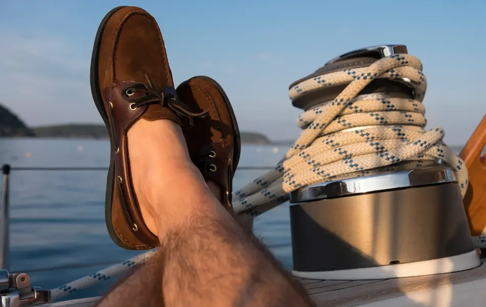 Manneben med brune seilersko lener seg på kanten av en seilbåt 