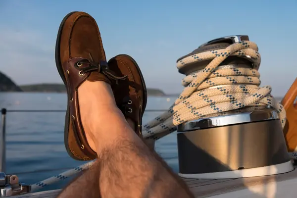 Manneben med brune seilersko lener seg på kanten av en seilbåt 
