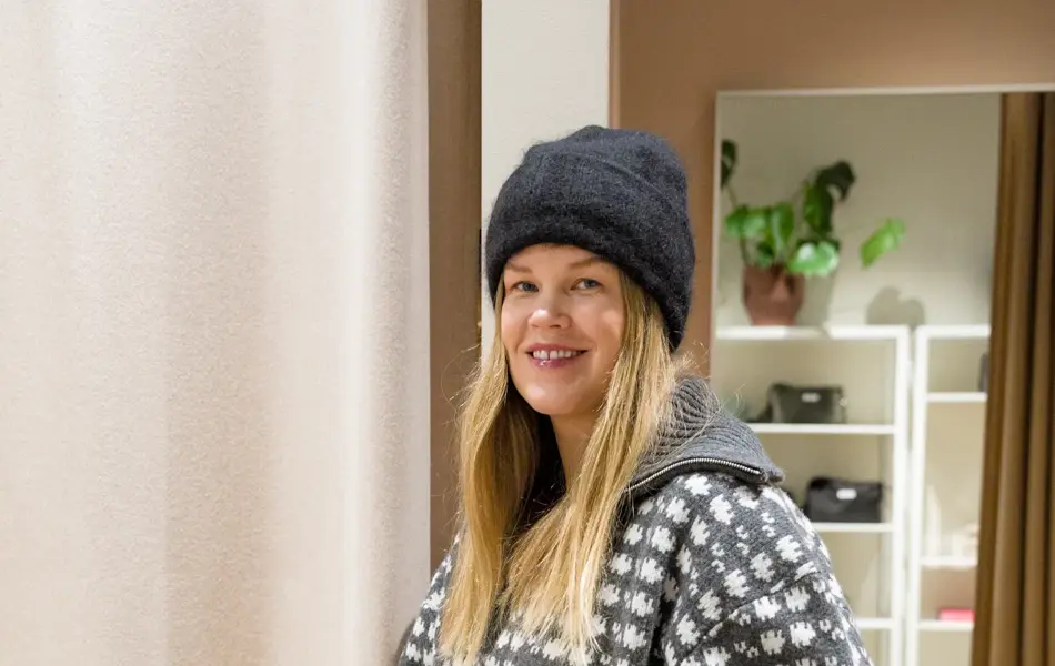 Dame med sort lue, mønstret jakke, sorte bukser og holder en rutete veske