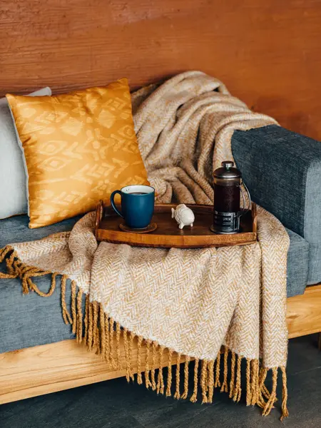 Trebrett med kaffekopp og presskanne står oppå gult ullpledd i blå sofa med gul pute 