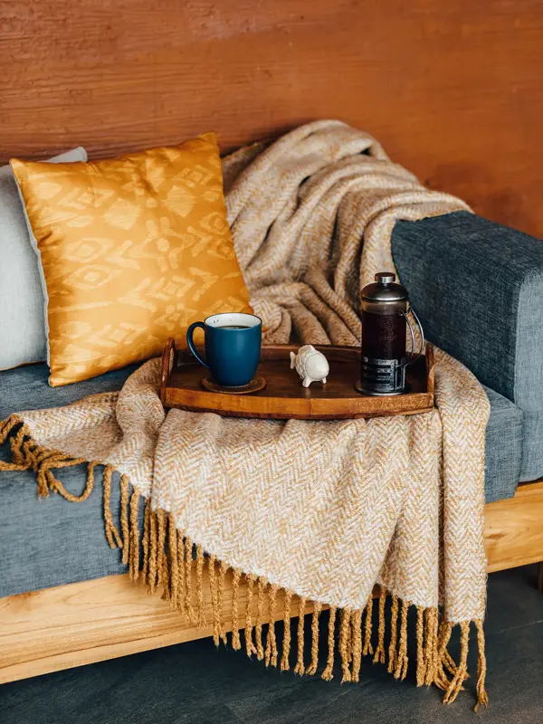 Trebrett med kaffekopp og presskanne står oppå gult ullpledd i blå sofa med gul pute 