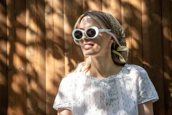 Portrait of a pretty girl in glasses and a headscarf standing against a wooden fence in the backyard on a Sunny day. High quality photo