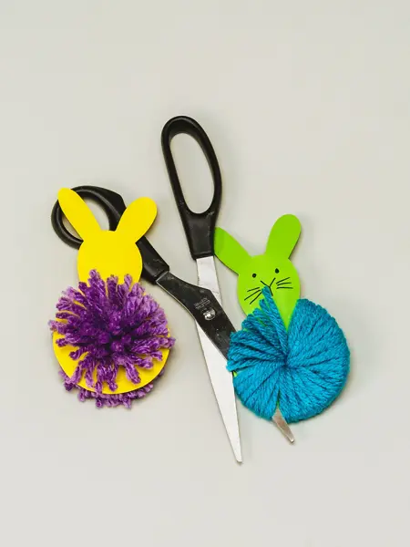 En gul og en grønn kanin laget av papir og garn ved siden av en saks 