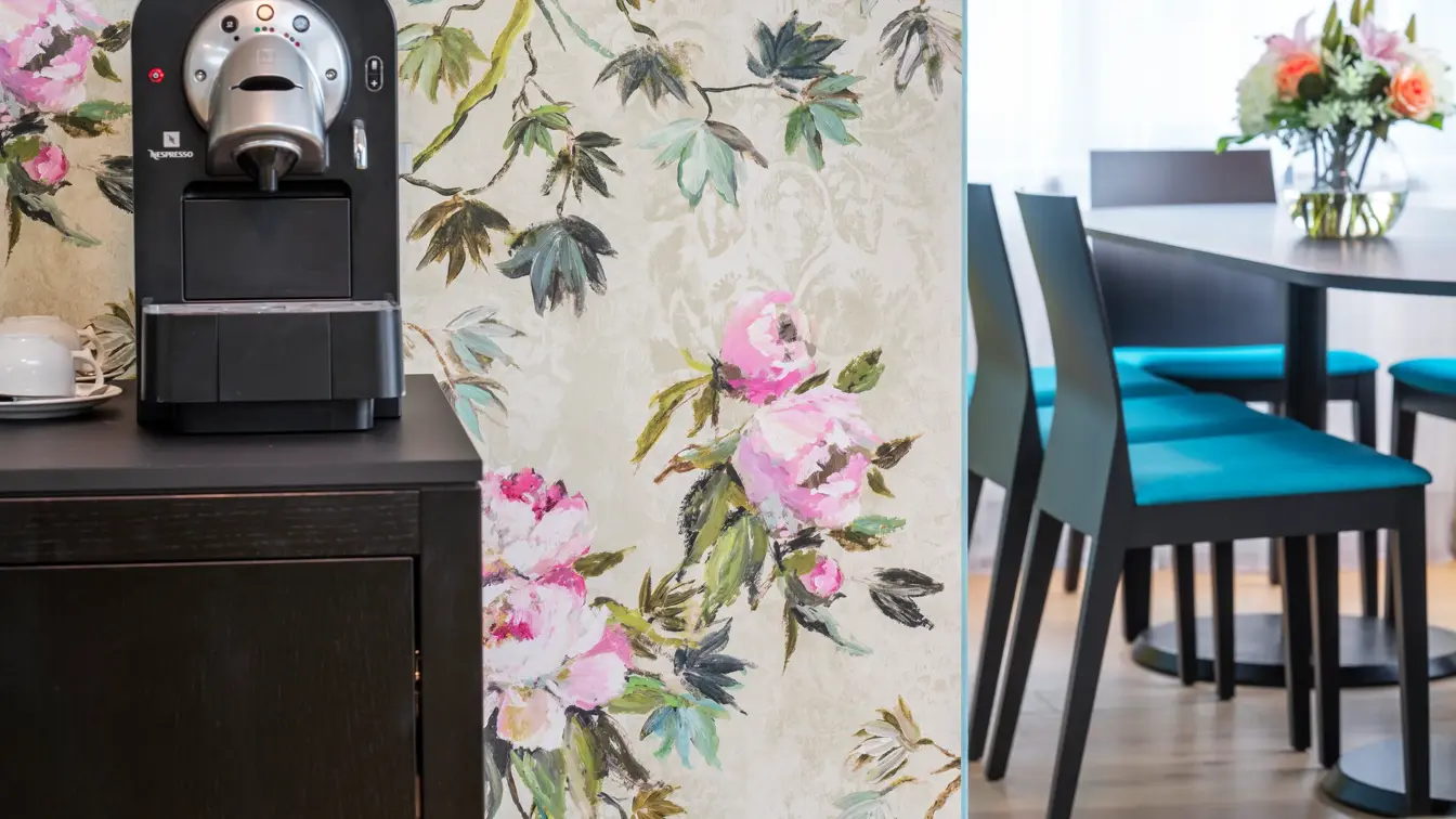 Blomstrete tapetvegg, kaffemaskin, spisebord med stoler. 