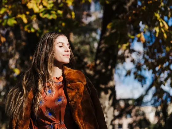 Dame med brunt hår, brun fuskepelskåpe og brun og blå silkebluse lukker øynene i solen foran høsttrær
