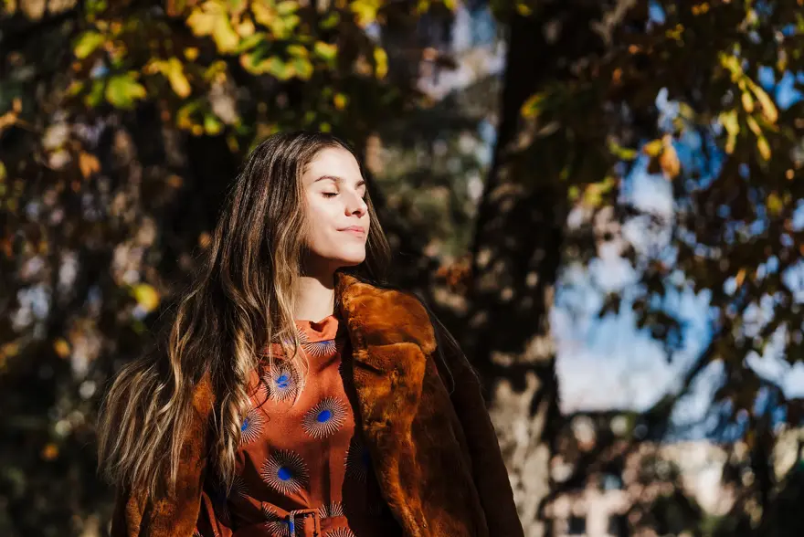 Dame med brunt hår, brun fuskepelskåpe og brun og blå silkebluse lukker øynene i solen foran høsttrær