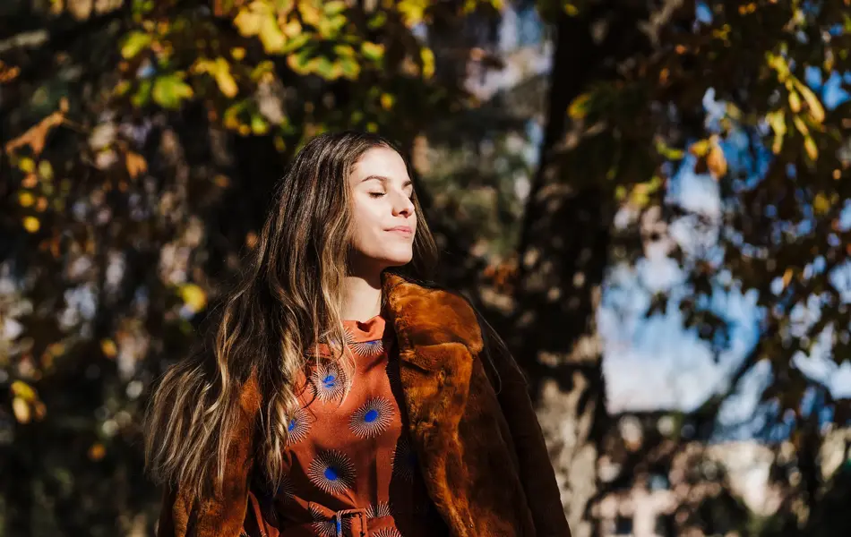 Dame med brunt hår, brun fuskepelskåpe og brun og blå silkebluse lukker øynene i solen foran høsttrær