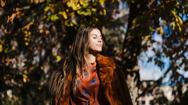 Dame med brunt hår, brun fuskepelskåpe og brun og blå silkebluse lukker øynene i solen foran høsttrær