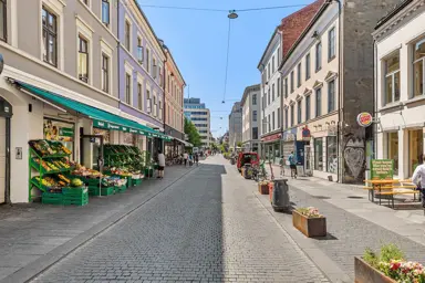 Handlegaten Torggata, sett fra øst til vest. Gågaten er pyntet med blomsterkasser og brostein. En grønnsakshandel på venstre side og restauranter med uteservering på høyre side.