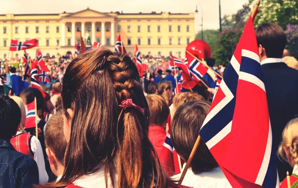 Mennesker står utenfor slottet med norske flagg på 17.mai, med fokus på jente med brunt hår med flette
