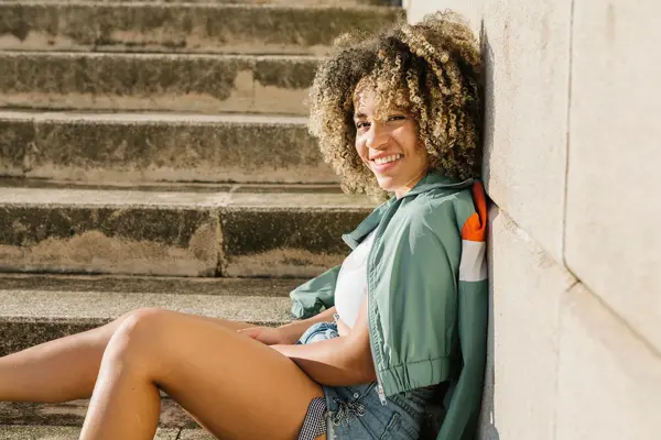 Dame i jeans-shorts og grønn treningsjakke sitter ute i en trapp og smiler til kamera