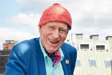 Nærbilde av Olav Thon, med signatur-luen, med en fin utsikt av Oslo bak