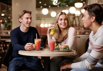 To gutter og en jente som sitter ved et bord i en kafé med drikke og mat på bordet