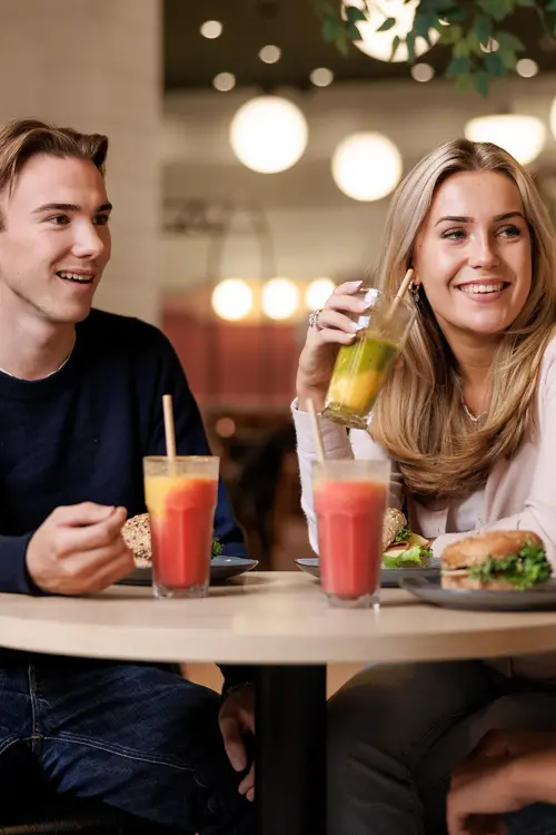 To gutter og en jente som sitter ved et bord i en kafé med drikke og mat på bordet