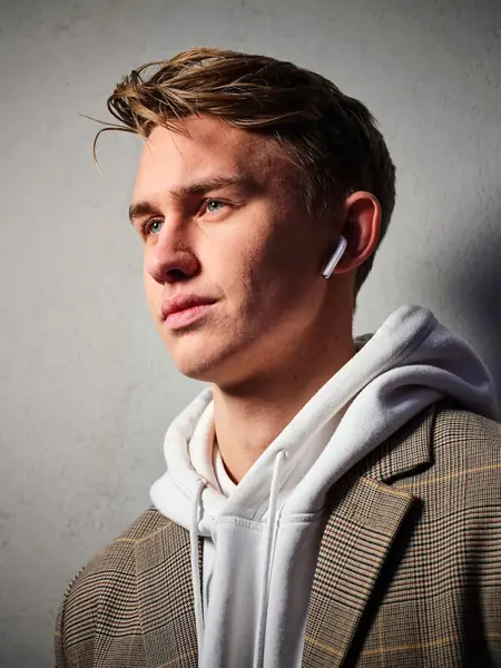 Ung mann iført hettegenser og jakke som står foran grå vegg og hører på musikk i Apple AirPods