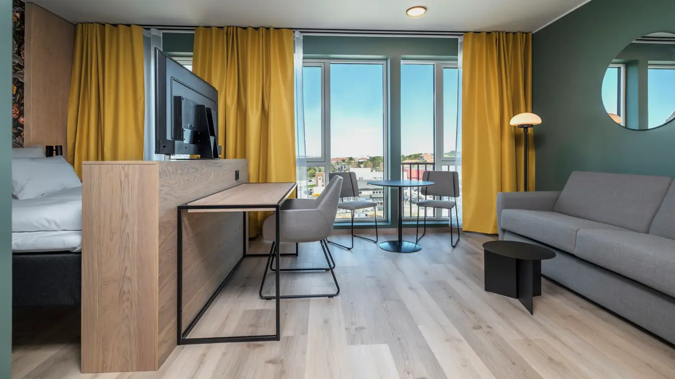 Superior rom med dobbeltseng, skrivebord med tilhørende stol, grå stol, grønne vegger, store vinduer og gule gardiner.