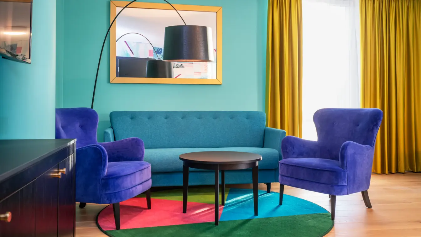 Turkis tapet, speil med gull ramme, gule gardiner, sittegruppe med turkis sofa, blå stoler og lite mørkt bord på fargerik teppe i business handicap room på Thon Hotel Storo i Oslo. 