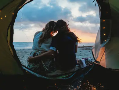 Young couple in love hug each other on the deserted beach on a summer evening at sunset during camping. Point of view inside the tent