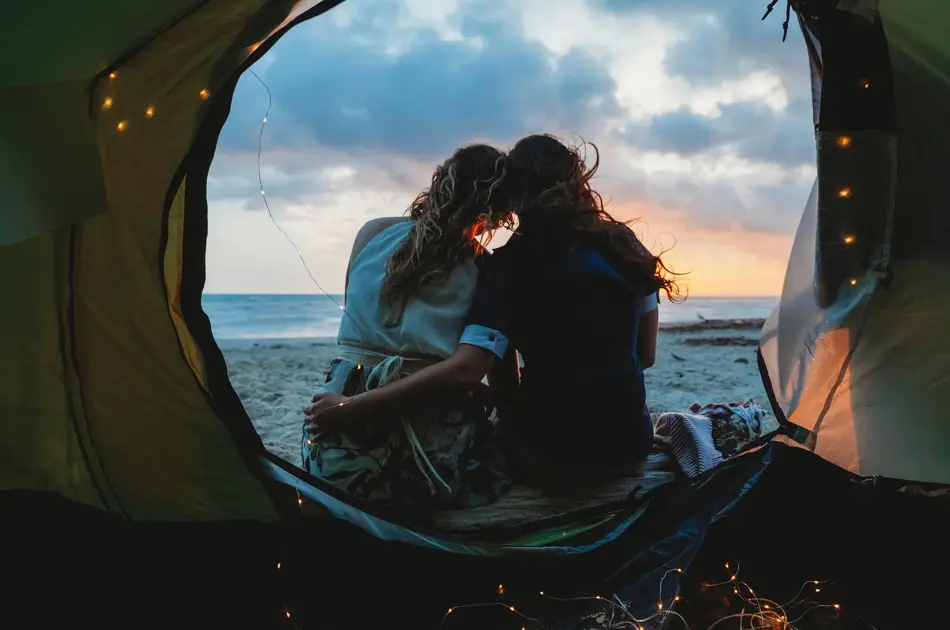 Young couple in love hug each other on the deserted beach on a summer evening at sunset during camping. Point of view inside the tent