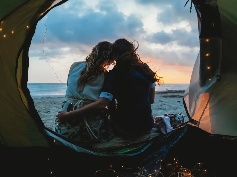 Young couple in love hug each other on the deserted beach on a summer evening at sunset during camping. Point of view inside the tent