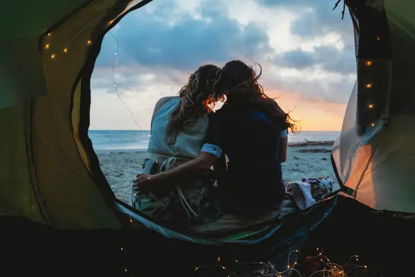 Young couple in love hug each other on the deserted beach on a summer evening at sunset during camping. Point of view inside the tent