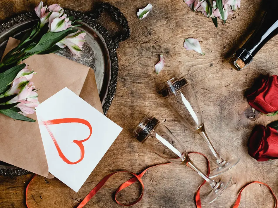 Blomster, to vinglass, kort med hjerte på og flaske på et bord