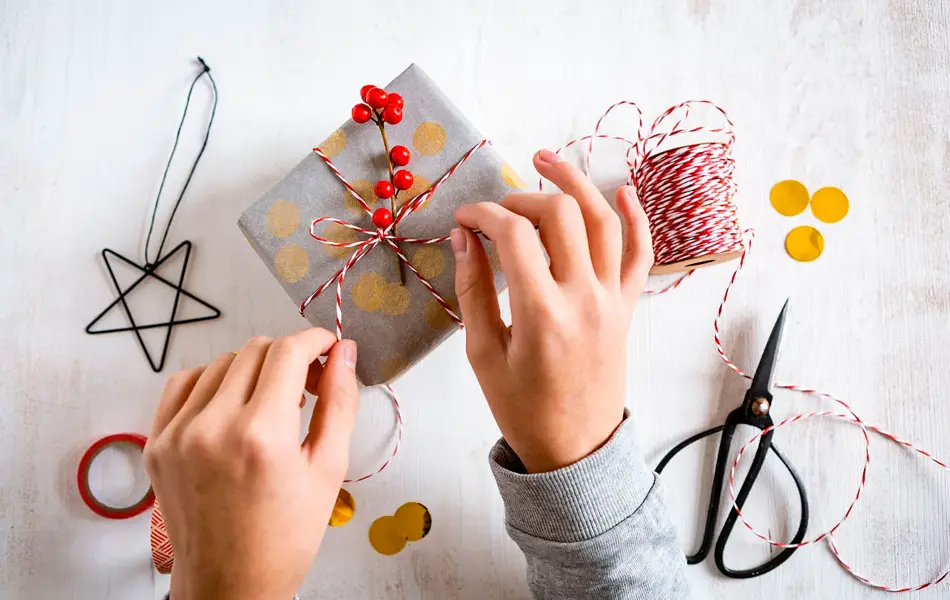 Bord med julegave, tilbehør til innpakning og to hender, sett ovenfra