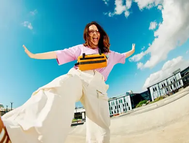 Dame med brune sko, hvit bukse og rosa topp smiler og sparker til kamera som ligger på bakken med blå himmel i bakgrunnen