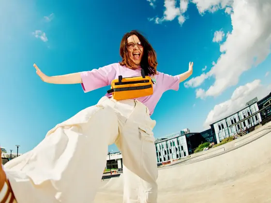 Dame med brune sko, hvit bukse og rosa topp smiler og sparker til kamera som ligger på bakken med blå himmel i bakgrunnen